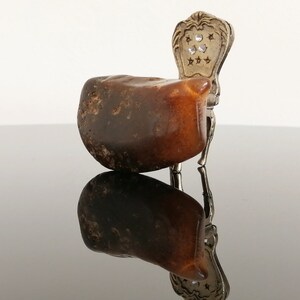 May include: A small, brown amber stone with a silver chair attached to it. The chair has a decorative back with a star pattern and clear rhinestones.