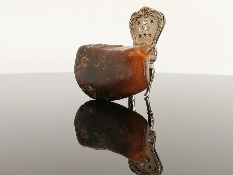 May include: A small, brown amber stone with a silver chair attached to it. The chair has a decorative back with a star pattern and clear rhinestones.