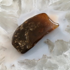 May include: A dark brown, irregularly shaped piece of amber on a white, textured surface.