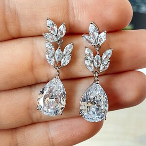 Può includere: Un paio di orecchini pendenti in argento con gocce di cristallo trasparente a forma di pera e un gruppo di cristalli trasparenti più piccoli in un design a foglia.