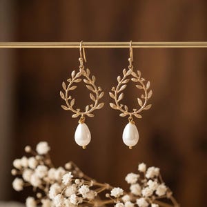 Peut inclure: Une paire de boucles d'oreilles pendantes dorées. Chaque boucle d'oreille présente un motif de couronne de laurier avec un pendentif en perle blanche en forme de goutte. Les boucles d'oreilles sont présentées sur un fond marron flou, avec des fleurs blanches au premier plan.