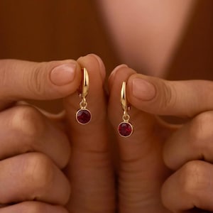May include: A pair of gold hoop earrings, each featuring a small, round, deep red gemstone charm suspended from the bottom. The earrings have a polished gold finish and a simple, circular design that holds the vibrant red stone securely in place.