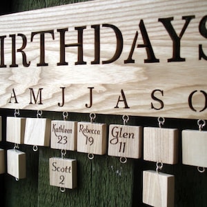 May include: A wooden birthday board with the word "BIRTHDAYS" engraved at the top. The board has slots for each month of the year, with small wooden tags hanging from each slot. The tags are engraved with names and numbers, such as "Kathleen 23", "Rebecca 19", and "Glen 11".