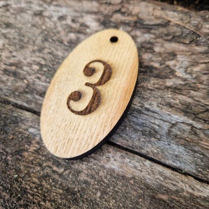 Puede incluir: Una etiqueta de madera de forma ovalada con el número 3 grabado en ella. La etiqueta tiene un agujero en la parte superior para colgarla.