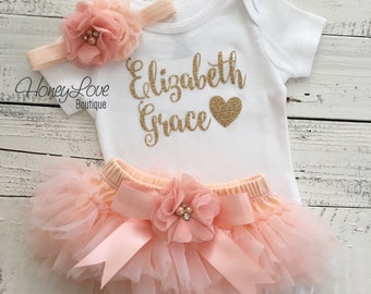 May include: Peach-colored baby outfit with a white bodysuit, tulle skirt, and headband. The bodysuit has gold glitter lettering that says "Elizabeth Grace" with a gold glitter heart.