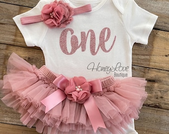 May include: A white baby bodysuit with the word "one" in glittery pink script, paired with a matching pink tutu skirt and headband. The tutu has multiple layers of tulle and a large bow. The set is designed for a baby's first birthday.