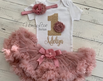 May include: A white onesie with a gold glitter number one and the name "Adelyn" in gold glitter. The onesie is paired with a dusty rose tulle tutu and a dusty rose headband with a flower.