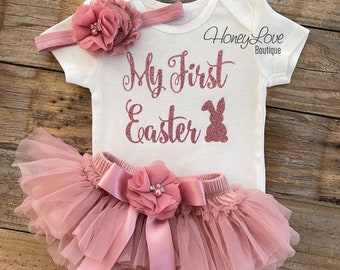 May include: A baby girl's Easter outfit in pink. The outfit includes a white bodysuit with the text "My First Easter" in pink glitter, a pink tulle tutu with a pink satin bow and a pink flower, and a pink headband with a pink flower.