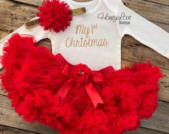May include: A white long-sleeved bodysuit with gold glitter text that reads "My 1st Christmas". The bodysuit is paired with a red tulle tutu with a red satin bow and a red flower with a rhinestone center. A red flower headband with gold glitter trim completes the outfit.