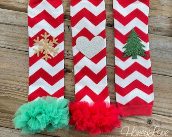 Op de afbeelding: Drie paar rode en witte chevron-beenwarmers met verschillende versieringen. Het eerste paar heeft een gouden sneeuwvlok, het tweede paar heeft een zilveren hart en het derde paar heeft een groene glitter-kerstboom. Elk paar heeft een tule-ruche aan de onderkant in een andere kleur.