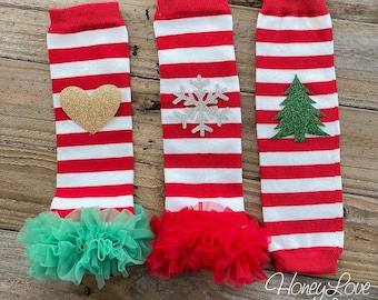 Op de afbeelding: Drie paar rood-wit gestreepte beenwarmers met verschillende feestelijke versieringen. De beenwarmers hebben een groene, rode en gouden tule ruche aan de onderkant. De versieringen zijn een gouden hart, een zilveren sneeuwvlok en een groene kerstboom.