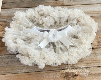 May include: A white tulle tutu with gold glitter dots and a white satin bow with a pearl embellishment. The tutu is on a wooden background.