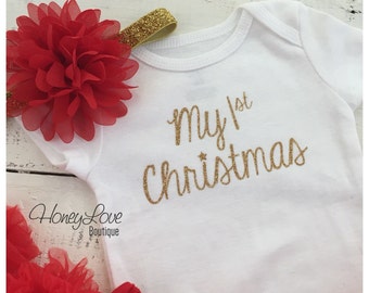May include: A white onesie with gold glitter lettering that says "My 1st Christmas". The onesie is paired with a red tulle skirt and a red flower headband with gold glitter trim.