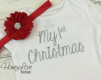 May include: White baby onesie with silver glitter text that reads "My 1st Christmas". A red flower headband with a rhinestone center is on top of the onesie.