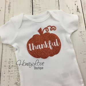 May include: White baby onesie with a copper glitter pumpkin design that says "thankful" on it.