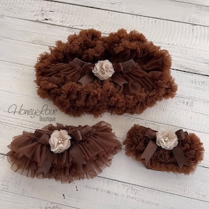 May include: Three brown tulle baby bloomers with a satin bow and a beige flower accent. The bloomers are layered and have a ruffled edge. The text "Honey Love Boutique" is visible in the image.