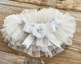 May include: White tulle baby bloomers with a white satin bow and a white flower with a pearl embellishment. The tulle has a subtle glitter pattern.
