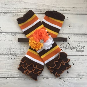 Puede incluir: Un par de calentadores de piernas a rayas marrones, naranjas y blancas con volantes marrones. Una diadema marrón con una gran flor naranja, amarilla y blanca está encima de los calentadores de piernas. El texto "Honey Love Boutique" está en la esquina inferior derecha de la imagen.
