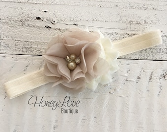 May include: A cream-colored baby headband with a large, layered flower in shades of beige and ivory. The flower is adorned with faux pearls and rhinestones. The elastic band is a matching cream color. The text "Honey Love Boutique" is printed on the bottom.