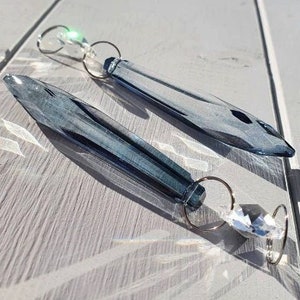 Op de afbeelding: Twee blauwe kristallen prisma zonnevangers met zilverkleurige ringen. Eén heeft een helder kristal, de andere een groen kristal. De langwerpige, facetgeslepen prisma's reflecteren licht. Ze liggen op een lichtgekleurd houten oppervlak.