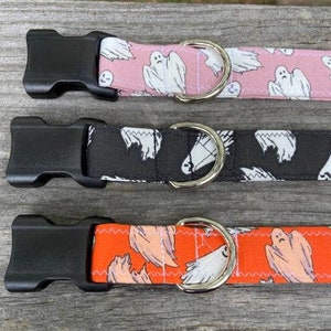 May include: Three pet collars featuring a ghost pattern. The top collar is pink, the middle is black, and the bottom is orange. Each collar includes a black plastic side-release buckle and a silver-toned metal D-ring for attaching a leash or tag.