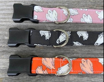 May include: Three pet collars featuring a ghost pattern. The top collar is pink, the middle is black, and the bottom is orange. Each collar includes a black plastic side-release buckle and a silver-toned metal D-ring for attaching a leash or tag.