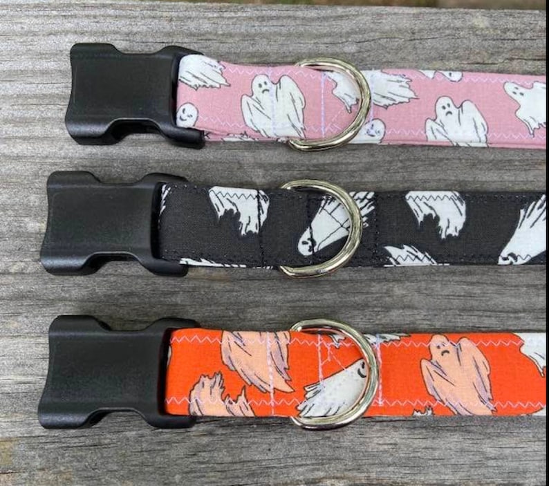May include: Three pet collars featuring a ghost pattern. The top collar is pink, the middle is black, and the bottom is orange. Each collar includes a black plastic side-release buckle and a silver-toned metal D-ring for attaching a leash or tag.