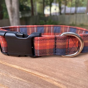 May include: A plaid dog collar in shades of red, orange, and navy blue. The collar features a black plastic buckle and a silver metal D-ring for leash attachment. The fabric has a classic plaid pattern.