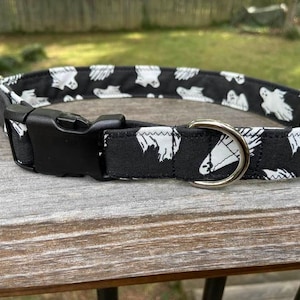 May include: A black fabric pet collar featuring a repeating white ghost pattern. The collar includes a black plastic side-release buckle and a silver-toned metal D-ring for attaching a leash. The design is consistent throughout the entire band.