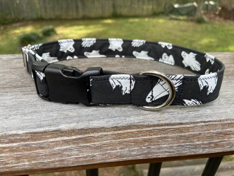May include: A black fabric pet collar featuring a repeating white ghost pattern. The collar includes a black plastic side-release buckle and a silver-toned metal D-ring for attaching a leash. The design is consistent throughout the entire band.