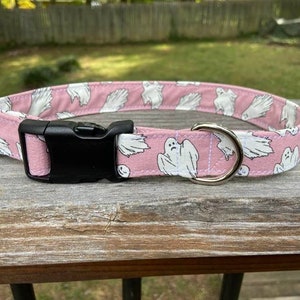 May include: A pink pet collar featuring a repeating pattern of white ghosts with black facial features. The collar includes a black plastic side-release buckle, a silver-toned metal D-ring for leash attachment, and a silver-toned metal adjustment slider.