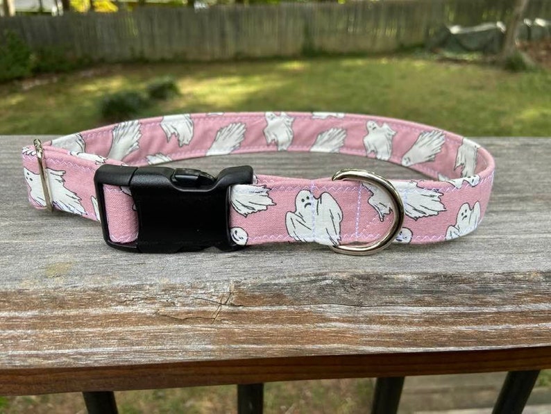 May include: A pink pet collar featuring a repeating pattern of white ghosts with black facial features. The collar includes a black plastic side-release buckle, a silver-toned metal D-ring for leash attachment, and a silver-toned metal adjustment slider.