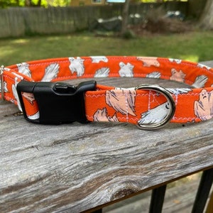 May include: An orange fabric pet collar featuring a repeating pattern of white ghosts and thin spiderwebs. The collar includes a black plastic side-release buckle, a silver-toned metal D-ring for leash attachment, and a silver-toned metal slider.