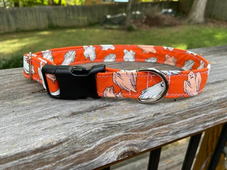 May include: An orange fabric pet collar featuring a repeating pattern of white ghosts and thin spiderwebs. The collar includes a black plastic side-release buckle, a silver-toned metal D-ring for leash attachment, and a silver-toned metal slider.
