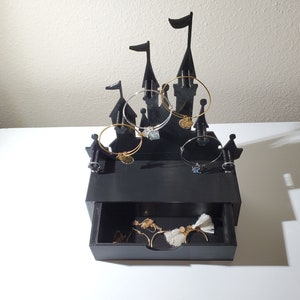May include: Black castle-shaped jewelry organizer with a drawer. The organizer has five hooks for bracelets and a drawer for storing other jewelry.