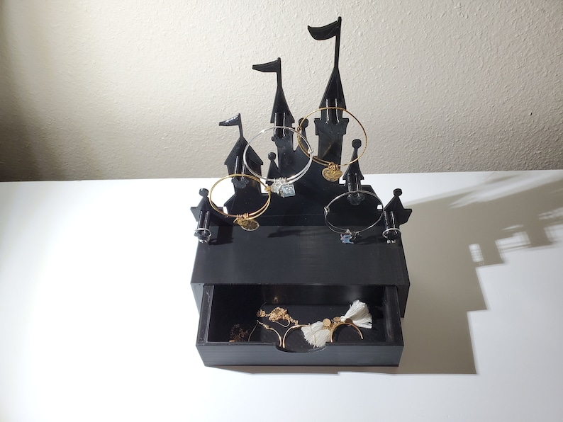 May include: Black castle-shaped jewelry organizer with a drawer. The organizer has five hooks for bracelets and a drawer for storing other jewelry.