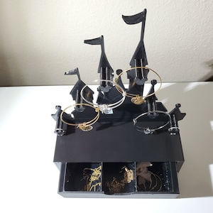 May include: Black castle-shaped jewelry organizer with a drawer. The top of the organizer has five hooks for hanging bracelets. The drawer has three compartments for storing smaller jewelry.