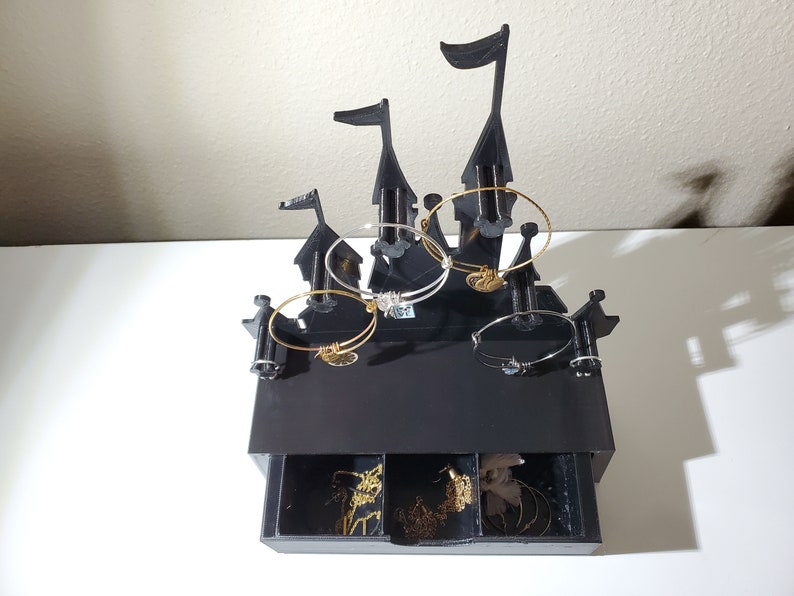 May include: Black castle-shaped jewelry organizer with a drawer. The top of the organizer has five hooks for hanging bracelets. The drawer has three compartments for storing smaller jewelry.