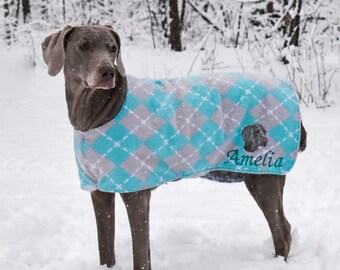 Puede incluir: Un perro Weimaraner gris con un suéter de rombos azul y gris con el nombre "Amelia" bordado. El perro está de pie en la nieve.