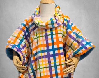 Puede incluir: Un poncho de forro polar a cuadros de colores con cuello de tortuga. El poncho es blanco con rayas naranjas, azules, moradas y amarillas.