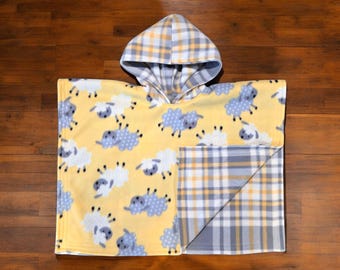 Puede incluir: Un poncho de forro polar amarillo con capucha, con un estampado de ovejas blancas y azules. La capucha y una esquina del poncho son a cuadros con cuadrados grises, amarillos y blancos. El poncho está diseñado para niños.