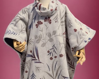 Puede incluir: Un poncho de forro polar gris con un estampado floral en tonos burdeos, azul y gris. El poncho tiene cuello de tortuga y se muestra en un maniquí con botas marrones.