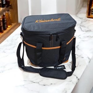 May include: Black insulated cooler bag with a brown leather trim and a zipper closure. The bag has a shoulder strap and the text "Kleinmachlmit" is embroidered on the front.