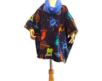 Puede incluir: Un poncho de forro polar azul oscuro con un estampado de monstruos coloridos. El poncho tiene un cuello de capucha azul y se muestra en un maniquí de madera.