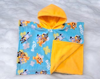 Puede incluir: Una manta con capucha azul y amarilla con estampado de perro de dibujos animados. La manta presenta las palabras "Bluey" y "Blue" repetidas en todo el estampado.