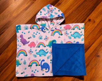 Puede incluir: Una manta de forro polar con capucha con estampado de dinosaurios y arcoíris, forrada con forro polar azul. La manta está doblada por la mitad y colocada sobre una superficie de madera.