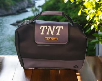 Puede incluir: Una bolsa de almuerzo aislada de color negro y morado con cierre de cremallera. La bolsa tiene un logotipo bordado en blanco que dice "TNT KANGA".