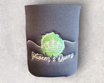 Puede incluir: Enfriador de latas de neopreno negro con un logotipo bordado en verde y blanco que dice "Between 2 Queens" y una corona.
