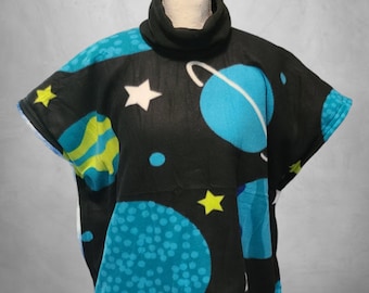 Puede incluir: Un poncho de forro polar negro con un estampado de temática espacial que presenta planetas, estrellas y un fondo de galaxia. El poncho tiene un cuello de tortuga y mangas cortas.