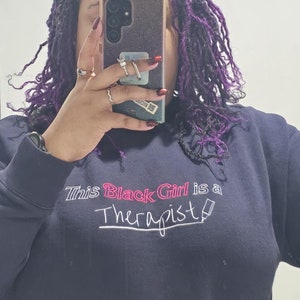 May include: A navy blue sweatshirt with the text "This Black Girl is a Therapist" embroidered in pink and white.
